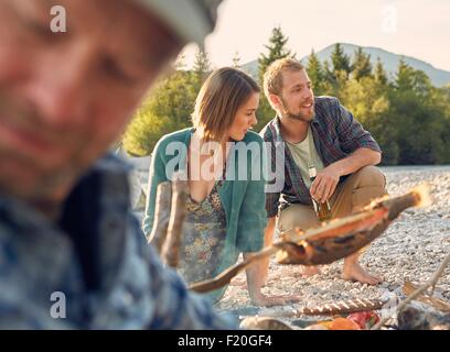 Young couple next to campfire, cooking fish, looking away Stock Photo