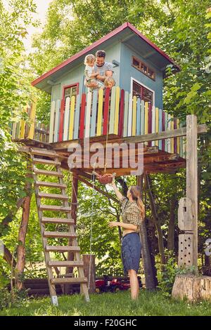Mother sending up basket of snacks to father and two sons, in tree house Stock Photo