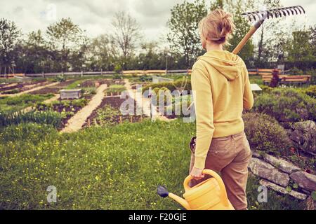 Mature woman, gardening, carrying rake and watering can, rear view Stock Photo