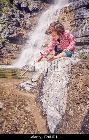 Young sitting on rock beside waterfall, holding stick, looking into water Stock Photo