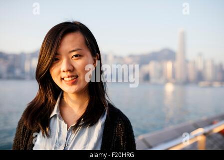 Portrait of young woman with long hair wearing shirt and cardigan in front of water, looking at camera