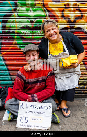 A Local Woman Poses With A Young Man Holding A Sign Asking If Passers By Would Like To Punch or Kick Him For £5, London, England Stock Photo