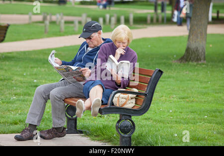 Middle aged couple relaxing, the lady with a paperback and the man with a newspaper on a park seat, England, UK Stock Photo