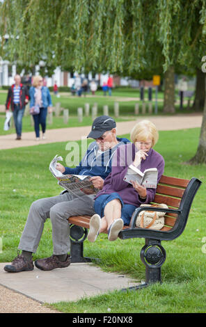 Middle aged couple relaxing, the lady with a paperback and the man with a newspaper on a park seat, England, UK Stock Photo