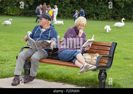 Middle aged couple relaxing, the lady with a paperback and the man with a newspaper on a park seat, England, UK Stock Photo
