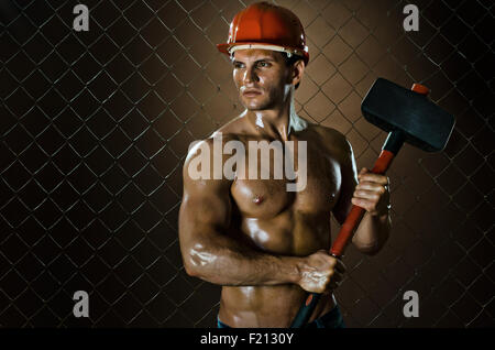 muscular worker , in  safety helmet  with big tup  in hands, on netting fence background Stock Photo