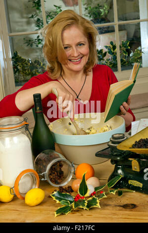 A woman in red mixing a Christmas pudding in a large bowl, using a recipe book and kitchen scales, with the ingredients. UK Stock Photo