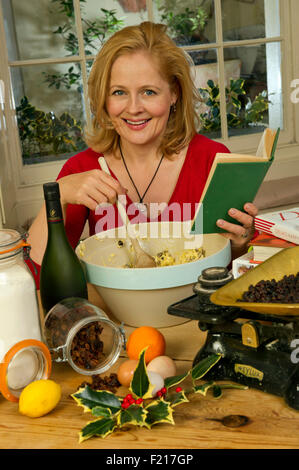 A woman in red mixing a Christmas pudding in a large bowl, using a recipe book and kitchen scales, with the ingredients. UK Stock Photo