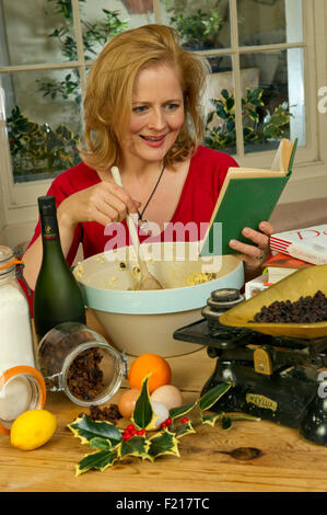 A woman in red mixing a Christmas pudding in a large bowl, using a recipe book and kitchen scales, with the ingredients. UK Stock Photo