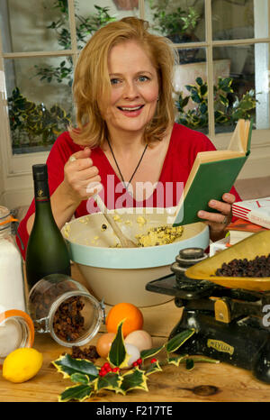 A woman in red mixing a Christmas pudding in a large bowl, using a recipe book and kitchen scales, with the ingredients. UK Stock Photo