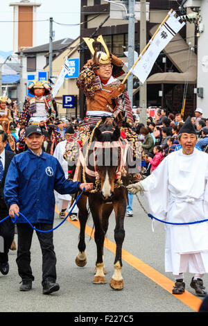 Genji festival parade in Japan. Man dressed in samurai full armour on horseback waving and smiling as he is lead through the  street by steward. Stock Photo