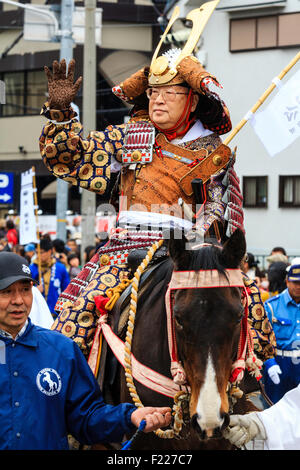 Genji festival parade in Japan. Man dressed in samurai full armour on horseback waving and smiling as he is lead through the  street by steward. Stock Photo