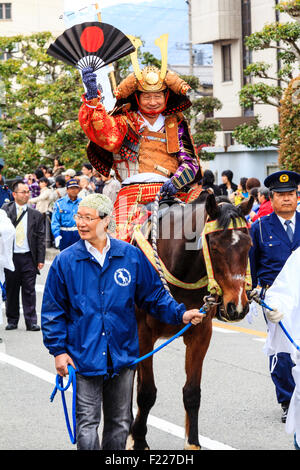 Genji festival parade in Japan. Man dressed in samurai full armour on horseback waving and smiling as he is lead through the  street by steward. Stock Photo