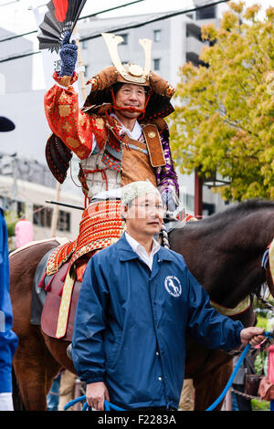 Genji festival parade in Japan. Man dressed in samurai full armour on horseback waving and smiling as he is lead through the  street by steward. Stock Photo