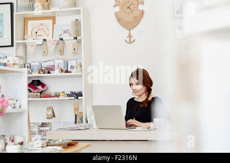 A woman sitting at a desk in a small gift shop, doing the paperwork, managing the business, using a laptop. Stock Photo