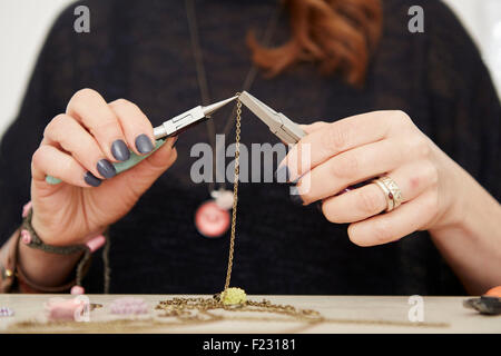 A woman seated at a workbench holding a gold chain with a small floral pendant, making jewellery. Stock Photo