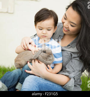 Smiling woman holding a rabbit, her young son sitting on her lap, stroking the animal. Stock Photo