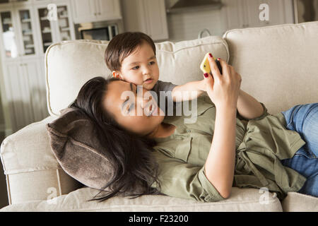 Woman lying on a sofa, smiling, cuddling with her young son and looking at a cell phone.