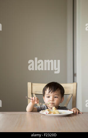 Young boy sitting on a chair at a table, eating a slice of cake. Stock Photo
