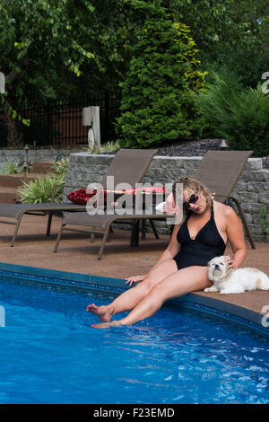 Trim Caucasian woman in her 50s sits on the edge of a residential swimming pool in her swimsuit petting her little white dog Stock Photo