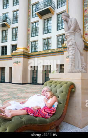 Caucasian senior woman and her granddaughter wearing a pink dress recline on a velvet chaise under a marble statue Stock Photo