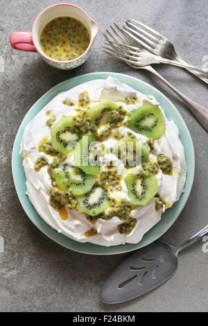 Pavlova with kiwi and passion fruit on a plate with a jug of passion fruit juice and cutlery over grey background.Top view Stock Photo