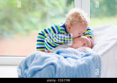 Cute little boy kissing his newborn brother. Toddler kid meeting new born sibling. Infant sleeping in white bouncer Stock Photo