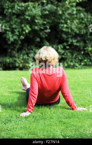 Back view of middle aged woman wearing coral pink coloured top and blue trousers sitting on grass outdoors Stock Photo
