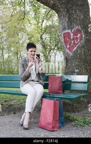 Mature woman on park bench applying lipstick for her date, Bavaria, Germany Stock Photo