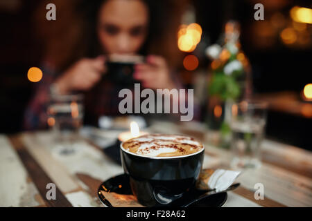 Close up shot of cup of coffee on table at cafe, with a woman drinking coffee in background. Focus on cup of coffee. Stock Photo