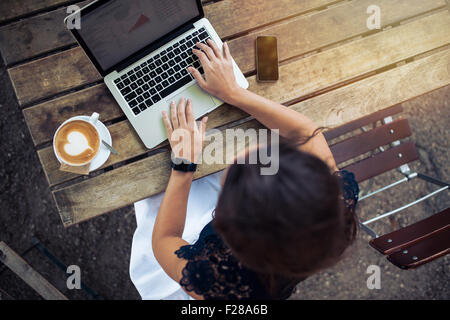 Top view of female using her laptop at a cafe. Overhead shot of young woman sitting at a table with a cup of coffee and mobile p Stock Photo