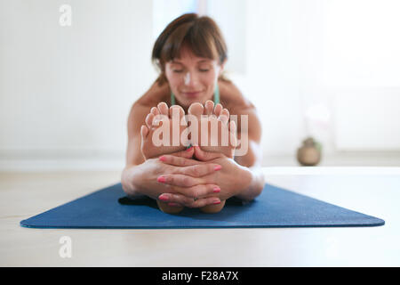 Woman sitting on exercise mat bending forward and holding legs. Woman practicing seated forward bend yoga exercise. Paschimotta Stock Photo
