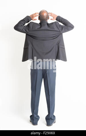 Rear view full body mature Indian business man hands scratching head , standing on plain background. Stock Photo