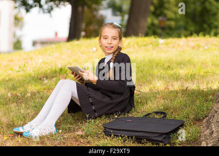 Small student girl sitting on grass and using tablet PC. Smiling pupil rest after school in park outdoors. Stock Photo