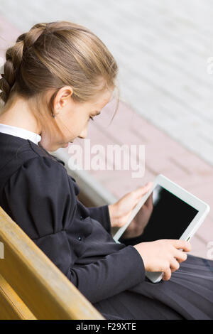 Small pupil girl with tablet sitting on bench. Schoolgirl plays with digital gadget Stock Photo