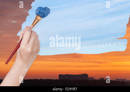 nature concept - seasons and weather changing: hand with paintbrush paints blue daily sky on orange sunset sky Stock Photo