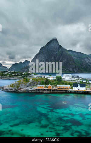 Mount Olstind above the yellow cabins and turquise waters of Sakrisoy fishing village on Lofoten islands in Norway Stock Photo