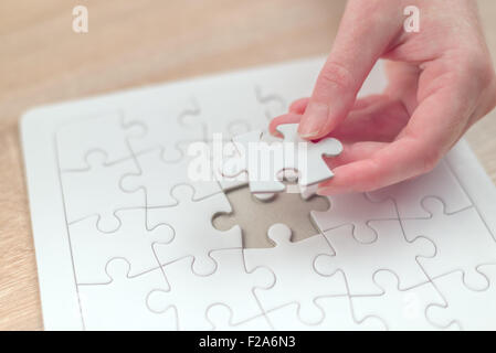 Female hand putting a missing piece and solving blank white jigsaw puzzle placed on top of old wooden oak table, close up Stock Photo