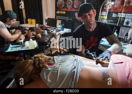 Garden City, New York, USA. September 13, 2015. R-L, JASON ACKERMAN and TROY RADECKI, a pro team of tattoo artists, are tattooing two young woman, lying prone, at the United Ink Flight 915 Tattoo convention at the Cradle of Aviation Museum in Long Island. Credit:  Ann E Parry/Alamy Live News Stock Photo