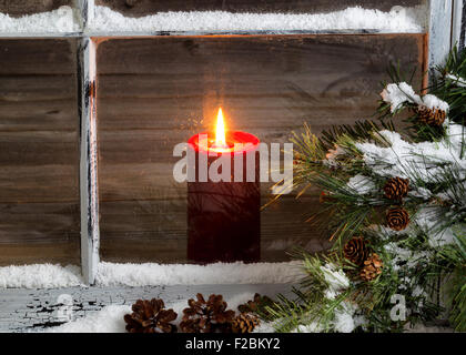 Decorated window with glowing red candle, selective focus on flame and top part of candle, pine tree, cones and snow outside. Stock Photo