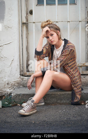Contemplative, teenage girl in shorts and a fashionable sweater resting on the back steps of a house in an alley. Stock Photo