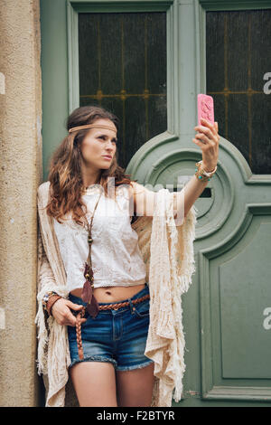Portrait of hippie woman in boho chic clothes standing outdoors against wooden door in old town and making selfie Stock Photo