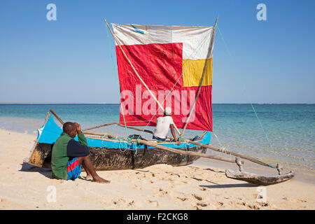 Two fishermen with traditional fishing pirogue with sail on beach at Nosy Ve, Madagascar, Southeast Africa Stock Photo