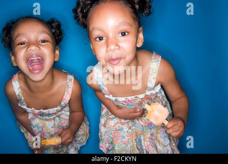 Two young identical twin girls eat orange popscicles in front of blue backdrop Stock Photo