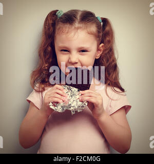 Cunning kid girl eating dark chocolate with pleasure and curious look. Vintage closeup portrait Stock Photo