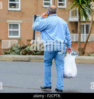 Man walking along a path carrying shopping and speaking on a mobile phone (cellular phone or cell phone) in England, UK. Stock Photo