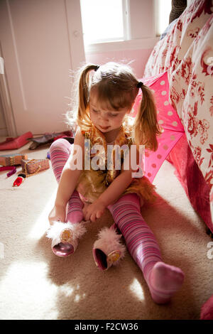 Young girl in pink plays sits on floor with bright light behind her, trying on shoe, playing dress-up in her bedroom Stock Photo