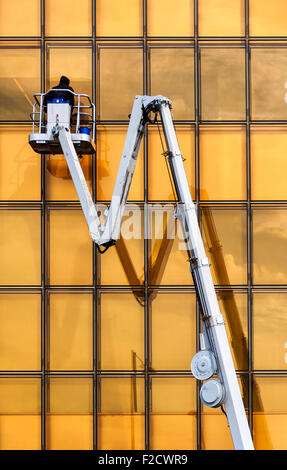 A window cleaner cleans the golden colored glass of a modern building from the safety of a mechanical crane. Stock Photo