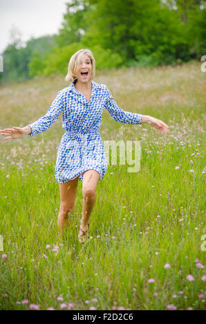 Girl with Blonde Hair Runs Through Field in Blue Dress while Laughing and Waving Her Arms Stock Photo