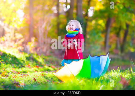 Little girl playing in the rain in autumn park. Child holding umbrella walking in the forest on a sunny fall day. Stock Photo